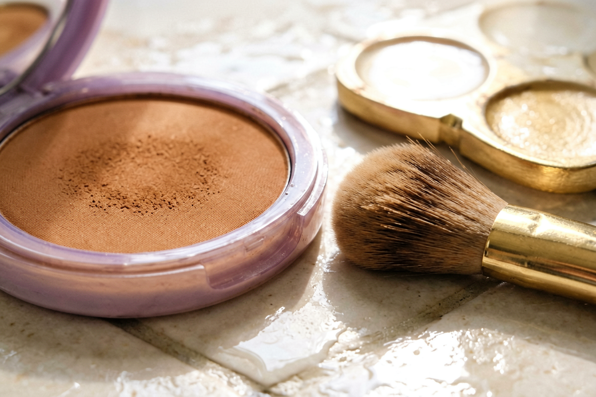 Editorial close-up macro shot. The edge of an unlabeled warm bronzer compact with visible matte powder texture beside so
